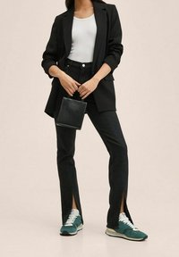 Veste noire portée sur un débardeur blanc, jean noir taille haute avec des fentes latérales, et baskets turquoise avec des accents blancs. Sac à main noir en main.