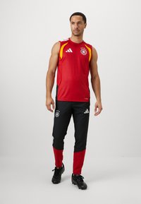 Piros ujjatlan mez textúrával és sárga részletekkel, fekete nadrággal kombinálva, amelynek alsó része piros és logó részletek találhatók rajta. Fekete cipők.