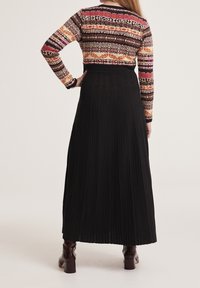 Dlouhá černá plisovaná sukně v kombinaci s multibarevným vzorovaným svetrem s dlouhými rukávy a přiléhavým pasem. Hnědé boty na podpatku.