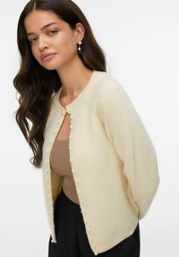 Cardigan crème en matière douce, avec des accents en perles le long des boutons avant et une coupe décontractée. Associé à un haut beige.