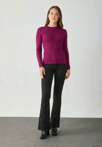 Blusa justa de mangas longas em fúcsia com padrão texturizado, combinada com jeans pretos flare e botas de tornozelo, contra um fundo neutro.