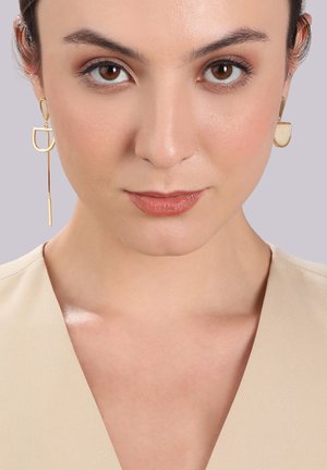 Gold-tone earrings featuring geometric shapes: a rounded stud and a long, thin drop. The texture is smooth and metallic.