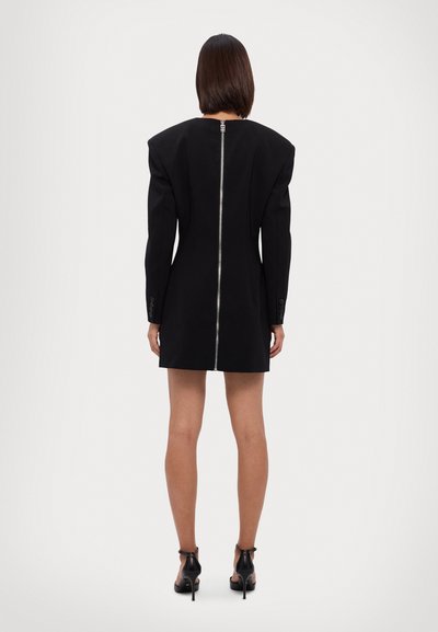 Robe noire structurée avec épaules padding, détail de fermeture éclair au dos et manches longues. Tissu lisse, longueur au-dessus du genou, portée avec des talons noirs.