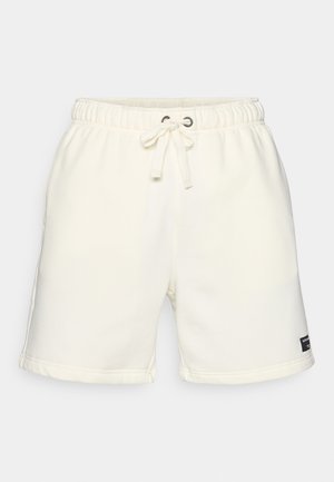 Light beige cotton shorts with an elastic waistband, drawstring closure, side pockets, and a small black label near the hem.