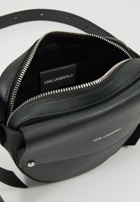 Zwarte leren crossbodytas met zilverkleurige hardware, voorzien van een ritssluiting, verstelbare band en een logotag aan de binnenkant. Glad oppervlak.