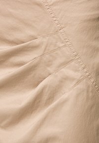 Beige tyg med en slät textur som har veck och sydda detaljer. Mönstret är subtilt och betonar tygets drapering och linjer.