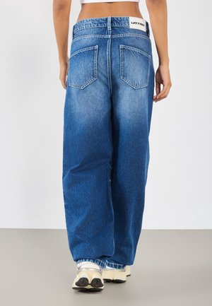 Personne portant un jean bleu ample à jambes larges et des baskets blanches, vue de dos sur un fond clair uni.