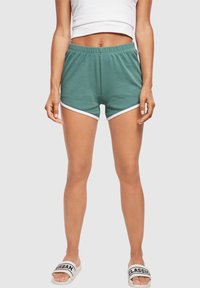 Woman wearing green shorts with white trim, white crop top, and white slide sandals with black text on a plain background.