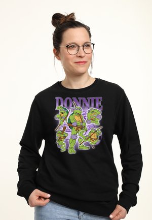 Kvinde iført briller og en sort sweatshirt med fem animerede Teenage Mutant Ninja Turtles-figurer og ordet "DONNIE."