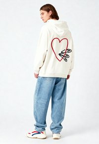 Felpa bianca con un grande cuore rosso e spille da balia nere ricamate sul retro, abbinata a jeans baggy di denim azzurro e scarpe da ginnastica bianche.