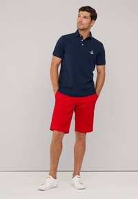Granatowa koszulka polo z trzema guzikami i haftowanym logo, w połączeniu z jaskrawoczerwonymi szortami. Wykończona białymi trampkami, prezentująca się na neutralnym tle.