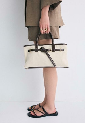 Femme portant une tenue marron et des sandales noires, tenant un sac à main beige et marron orné de garnitures en cuir et de détails de boucles dorées.