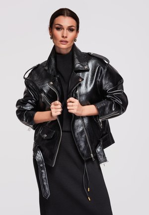 Kobieta trzymająca przednią część błyszczącej czarnej skórzanej kurtki, ubrana w czarną sukienkę i złote kołowe kolczyki, na jasnym, jednolitym tle.