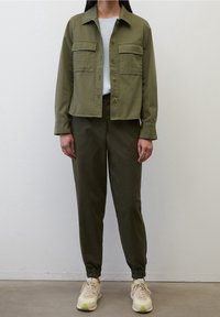 Chaqueta corta de color verde oliva con dos bolsillos en el pecho, combinada con pantalones holgados verdes oliva y zapatillas ligeras con acentos amarillos.
