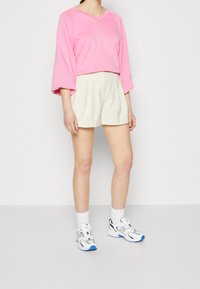 Jasnoróżowa krótka bluza z szerokimi rękawami, w zestawieniu z kremowymi szortami. Modelka ma na sobie białe sneakersy z niebieskimi i zielonymi akcentami.