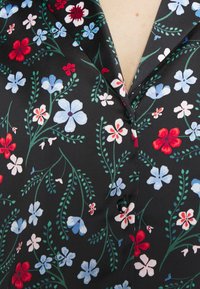 Blusa in tessuto con motivo floreale caratterizzata da fiori rossi, bianchi e blu su uno sfondo nero, con una texture liscia e chiusura a bottoni.