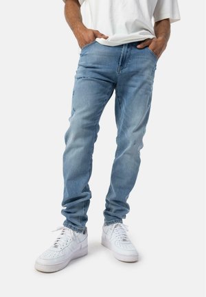Lyseblå slidte denimjeans med slim fit, flossede detaljer og lommer. Båret med hvide sneakers. Minimal synlig branding.