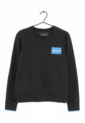 Czarna bluza crewneck z niebieską naszywką "Calvin Klein Jeans" na piersi i dopasowanym niebieskim obszyciem na mankietach, wisząca na czarnym wieszaku.