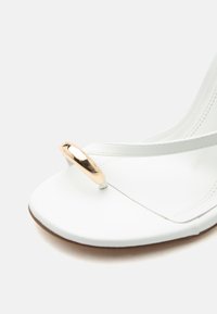 Λευκό σανδάλι ανοιχτού δαχτύλου με λεπτό λουράκι και χρυσή διακοσμητική λεπτομέρεια στη θέση του δακτύλου, τοποθετημένο πάνω σε ένα απλό ανοιχτό φόντο.