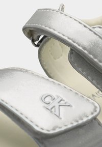 Εγγύς λήψη ασημένιων μεταλλικών λουριών σανδαλιών με αυτοκόλλητα βέλκρο και χαραγμένο λογότυπο "CK" σε ένα λουρί.