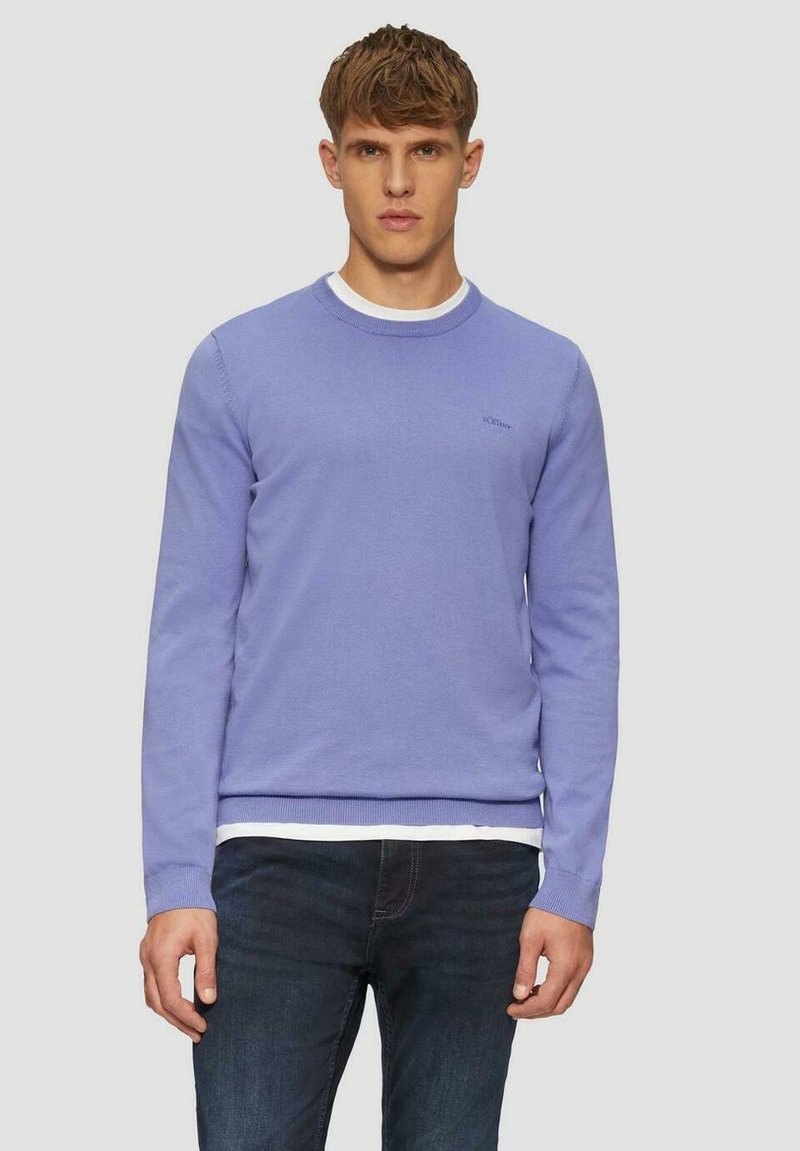 Lichtpaarse sweater met een ronde hals en geribde manchetten. Kenmerkt zich door een subtiele logo op de borst. G gedragen over een witte T-shirt en blauwe jeans.