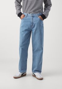 Person som bär ljusblå löst sittande jeans, en svartvit randig långärmad skjorta och vita sneakers med bruna sulor, händerna i fickorna.