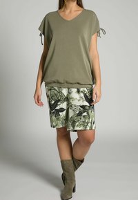 Mujer vestida con una blusa verde oliva de escote en V y mangas anudadas, pantalones cortos con estampado de hojas tropicales y botas de tacón alto de ante color topo sobre fondo gris.