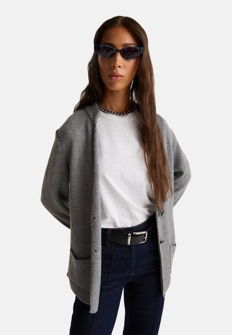 Cárdigan de punto gris con un patrón texturizado, sobre una camisa blanca. Jeans de mezclilla oscura y cinturón negro con hebilla plateada. Las gafas de sol completan el conjunto.