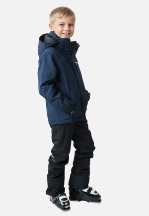 Granatowa wodoodporna kurtka z kapturem, zapinana na zamek, z regulowanymi mankietami, w połączeniu z czarnymi spodniami i czarnymi butami narciarskimi z akcentami.