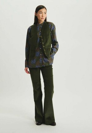 Veste en velours côtelé vert sur une chemise à motifs avec des designs géométriques bleus, verts et bruns, assortie à un pantalon en velours côtelé évasé.