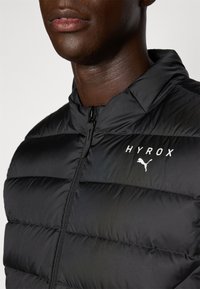 Veste en duvet noire avec un design matelassé horizontal, dotée d'une fermeture éclair, d'un col haut et d'accents en blanc "HYROX" et du logo Puma.