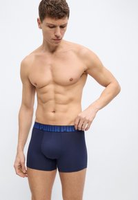 Νεαρός άνδρας χωρίς πουκάμισο που ρυθμίζει τη μέση της μπλε καθημερινής εσώρουχας Puma μπροστά σε απλό φωτεινό φόντο.