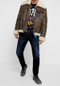Giacca in pelle marrone con colletto in shearling, maglione nero a motivo, jeans scuri e scarpe grigie. La giacca presenta borchie sulle spalle.