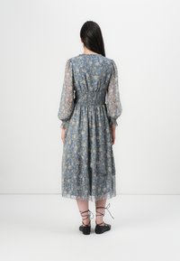 En ung kvinna står med ryggen mot kameran, klädd i en blå klänning med blommönster och svarta snörskor i en minimalistisk miljö.