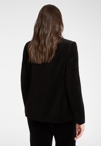 Blazer in velluto nero con maniche lunghe, taglio su misura e tessuto liscio. Presenta una cucitura verticale di dettaglio e un taglio dritto sul retro.