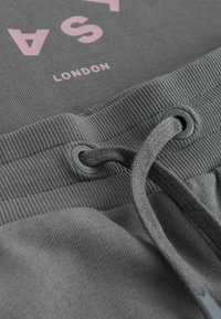 Detail sivého textilu s ružovým nápisom „LONDON“ a sivým sťahovacím šnúrkam prestrčeným cez očká na rebrovanom páse.