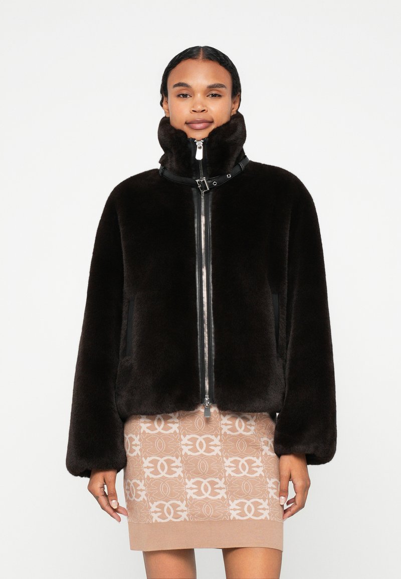 Casaco de pele sintética castanho com gola alta, fecho de correr à frente, detalhes pretos nos ombros e bolsos frontais, combinado com uma saia bege estampada.