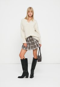 Maglione oversize beige chiaro con scollo a V, gonna mini a quadri in tonalità marroni, stivali neri al ginocchio con tacco a blocco e una piccola borsa scura.