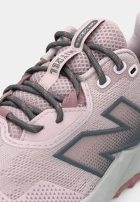 Gaiši rozā sporta apavi no tīkliņa ar pelēkām detaļām, teksturētu augšdaļu un kontrastējošām tumšām šņorēm. Uz sāniem redzams izteiksmīgs logotipa elements.