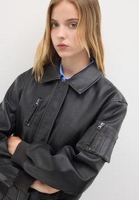 Giacca bomber in pelle nera con un ampio colletto, tasche con zip sul petto e sulla manica, e polsini a coste per una vestibilità aderente.