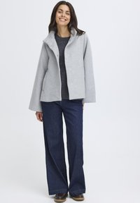 Grå jakke med høj krave og trykknapper, over en mørk sweater. Kombineret med vide blå jeans og brune støvler. Glat tekstur.