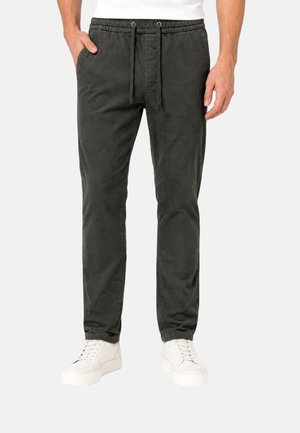 Man draagt donkergrijze corduroy joggerbroek met elastische tailleband en trekkoord, gecombineerd met witte sneakers op lichte achtergrond.