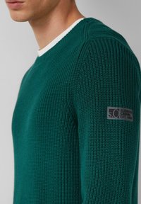 Zielony sweter z dzianiny o fakturowanym wzorze. Posiada biały okrągły dekolt oraz szary metkę na lewym rękawie z napisem "Elevated Casual Est. 1969."