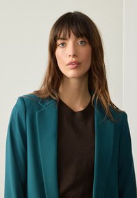 Teal blazer with a structured lapel over a brown sweater. The fabric appears smooth, with long, wavy hair framing the face.