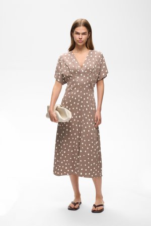 Mujer de pie con vestido midi marrón con lunares, sosteniendo un bolso blanco y llevando sandalias negras sobre un fondo blanco liso.