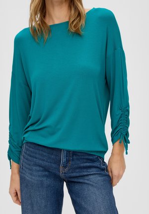 Femme portant un haut à manches longues bleu sarcelle avec manches froncées à cordon et un jean en denim bleu, debout avec une main reposant à côté.