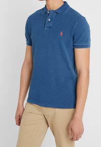 Man som bär en blå kortärmad polo med en röd broderad logotyp och beige byxor, stående mot en enfärgad bakgrund.