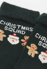 Dunkelgrüne Socken mit weicher Textur und gestrickten Motiven von Santa und Lebkuchenmännern, beschriftet mit "CHRISTMAS SQUAD" in Weiß.