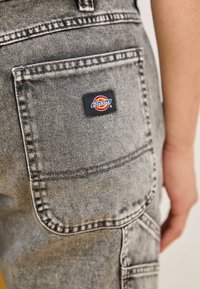 Sive traperice s naramenicama s velikim stražnjim džepom sa šivenim detaljima i crnom oznakom "Dickies". Tkanina pokazuje blagu teksturu.
