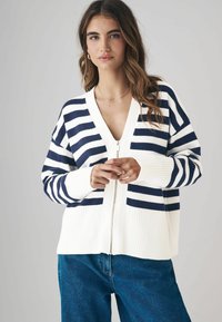 Cardigan a strisce con zip in blu navy e crema. Presenta polsini e orlo a costine. Indossato sopra pantaloni in denim, con scollatura a v e vestibilità rilassata.